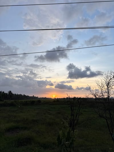 A vertical shot of the sky over Pulorungkom, Indonesia, displays a colorful sunset. The sky has blue sections interspersed with gray and white clouds. There are black power lines running vertically along the left side of the picture. The horizon is a thin line of orange and yellow where the sun is setting. The silhouettes of trees and vegetation are visible at the bottom of the image. Some bare tree branches are at the top right.