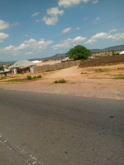 This image, taken in Kaltungo, Nigeria, depicts an outdoor scene under a clear blue sky with scattered white cumulus clouds. In the immediate foreground, a dark grey asphalt road, exhibiting minor wear and faded white road markings on its left side, traverses the lower portion of the frame. Beyond the road, a wide expanse of reddish-brown dirt and sand, characteristic of laterite soil, dominates the midground. Patches of green vegetation and small shrubs are interspersed within this unpaved area.

To the left of the unpaved area, a light-colored, single-story building with a greyish-green pyramidal roof is visible, featuring a small attached lean-to structure at its front. Adjacent to this building and extending towards the right, a low wall constructed from light brown concrete blocks encloses a property. A dirt path branches from the main unpaved area, leading towards the walled properties. Behind the wall, a prominent green tree with dense foliage stands. Further behind the wall, additional residential structures with light-colored roofs are partially visible.

In the background, a range of distant, low-lying hills or mountains with sparse vegetation lines the horizon. Several thin black power or communication lines stretch horizontally across the upper mid-section of the sky. No individuals, animals, or specific actions are discernible within the frame. The scene presents a static view of a semi-urban or rural environment under bright daylight conditions.