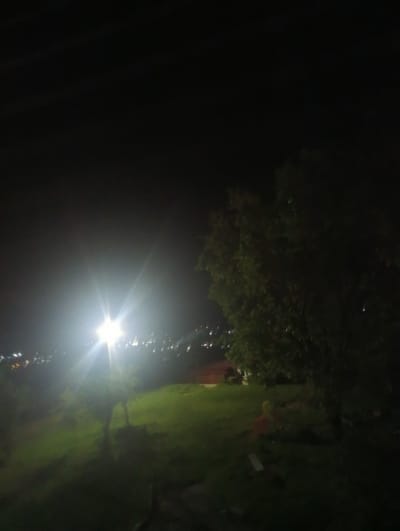 This is a nighttime photograph taken in the city of Rezina, Moldova. The image captures a dimly lit outdoor scene, with a bright artificial light source dominating the left side of the frame, casting a strong glare and illuminating the surrounding grassy area. In the distance, a faint glow suggests the presence of urban lights. A large tree with dark foliage is visible on the right, contributing to the overall shadowy atmosphere. The ground appears to be covered in grass and is uneven in texture. There are hints of structures or objects in the mid-ground, partially obscured by darkness. A solitary figure, possibly wearing a red garment and a yellow covering, is faintly visible in the lower right quadrant of the image, standing on the grass.