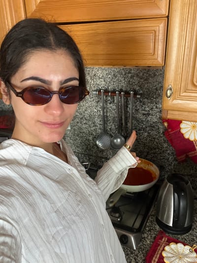 The image depicts a woman wearing sunglasses and a white striped shirt in a kitchen. She is partially visible in the foreground, with her face angled towards the camera. In the background, there are kitchen cabinets, cooking utensils hanging on a rack, a pot with a red sauce on a stovetop, a kettle, and a red hot pad. The countertop is made of a speckled gray material. The location cannot be determined to be Èze, France from the image alone.