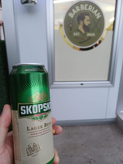 A human hand is prominently positioned in the foreground, holding a 500ml Skopska Pivara "SKOPSKO" Lager Beer can. The can is green and cream-colored, displaying text such as "THE ORIGINAL RECIPE," "PREMIUM QUALITY," "SINCE 1924," and "ПИВАРА СКОПЈЕ" (Brewery Skopje). The scene is located in Gevgelija, North Macedonia. In the background, a light-colored storefront door or window is visible, featuring a circular green and brown logo. This logo depicts a bearded man in profile, flanked by a straight razor and scissors, with the text "BARBERIAN" and "EST. 2023." The ground surface in the lower portion of the image consists of dark grey concrete, with a patch of green artificial turf visible to the far left.