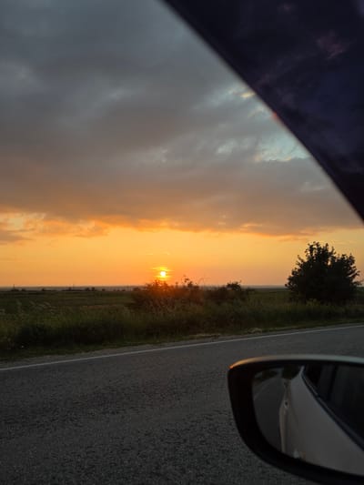 The image captures a serene sunset scene from the interior of a vehicle, looking out a side window, likely in Ivanovtsi, Bulgaria. The sky is dramatically colored with vibrant orange and yellow hues, indicative of dusk or dawn, though the low position of the sun on the right suggests sunset. The sun itself is visible as a bright, golden orb near the horizon. Scattered clouds traverse the sky, some catching the warm light, others appearing darker.

Below the colorful sky, a flat, rural landscape stretches into the distance, characterized by green fields, sparse trees, and bushes along the horizon. In the immediate foreground, a dark asphalt road with a distinct white line runs parallel to the vehicle, bordered by a strip of green, wild grass. A dark car window frame partially obscures the top and left edges of the view. To the upper right, the vehicle's side mirror is prominently visible, reflecting a distorted, darker view of the road and the car's body. No specific activity beyond being in or by a car is clearly depicted. There is no visible text in the media file.