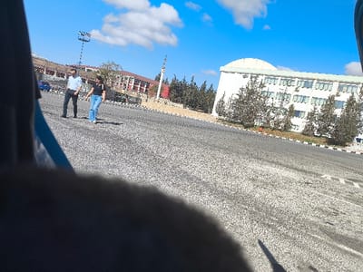 This outdoor, daytime image depicts a scene on the Near East University campus in Kızılbaş, Northern Cyprus / Cyprus, under a clear blue sky with scattered white cumulus clouds. The foreground is partially obscured by a dark, blurry, amorphous object, suggesting the photograph was taken from a vehicle or behind a barrier.
In the middle ground, two individuals are observed walking from left to right across a broad, textured dark grey asphalt surface. The individual on the left is a male, wearing a light blue collared shirt, dark trousers, and dark shoes. The individual on the right is a female, dressed in a dark t-shirt, light blue jeans, and white shoes. Both appear to be moving in the same direction.
The background features prominent structures and vegetation. To the right, a multi-story white building with a distinctive curved, light-colored roof is visible, characterized by numerous rectangular windows, many equipped with external air conditioning units. Several small, green coniferous trees are planted in front of this building. Further to the left and rear, a complex of reddish-brown, multi-story buildings with multiple windows is present. A tall pole featuring a square light fixture stands in the background, between the building complexes. A partial view of a decorative stone or concrete monument is visible in the mid-left background. A dark blue vehicle is partially visible behind the two walking individuals on the left. Shadows are cast towards the left and foreground, indicating the sun's position.