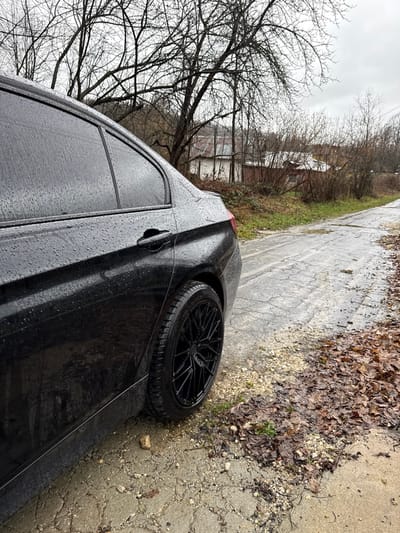 A dark-colored sedan, likely black, is parked on the right side of a narrow, wet, and cracked asphalt road in Peyovstsi, Bulgaria. The vehicle's rear driver's side, including the rear door, quarter panel, and a portion of the trunk lid, is visible. The car's exterior is covered in numerous water droplets, indicating damp conditions. A black multi-spoke aftermarket wheel with a low-profile tire is visible on the rear driver's side. The road surface is uneven, showing significant wear, cracks, and patches of light-colored gravel. To the immediate right of the road, a strip of ground is covered with damp, brown fallen leaves and sparse green vegetation. In the background, a dense stand of bare deciduous trees and bushes lines the road. Beyond the trees, several modest buildings are partially visible; one is light-colored with a dark roof, and another appears reddish-brown. The sky is overcast and gray. No individuals or active subjects are present.