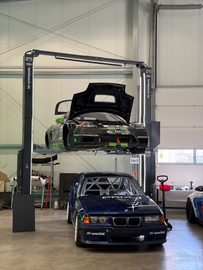 This image depicts an indoor automotive workshop facility in Meuspath, Germany. A black Twin Busch two-post car lift, marked with "4200KG" and "350 CE" specifications, occupies the central foreground. Elevated on this lift is a black and green Honda NSX JK race car, identified by the number "68," "JOHNDANBYRACING.com," and "HONDA" branding. Its engine hood is open, and both doors are slightly ajar. Orange straps are visible near the front undercarriage. Directly beneath the elevated vehicle, on the concrete floor, rests a dark blue BMW E36-series race car with a visible interior roll cage. This BMW features "N NANKANG" decals on its front bumper and a white, star-like graphic on its hood. The background consists of light-colored industrial paneled walls, a high ceiling with strip lighting, and a large white section or door labeled "Tor 2" (Gate 2). Various tools, equipment, and additional car parts, including a red stand and a power drill, are scattered on tables and shelves in the peripheral areas of the workshop. No individuals are present in the scene.