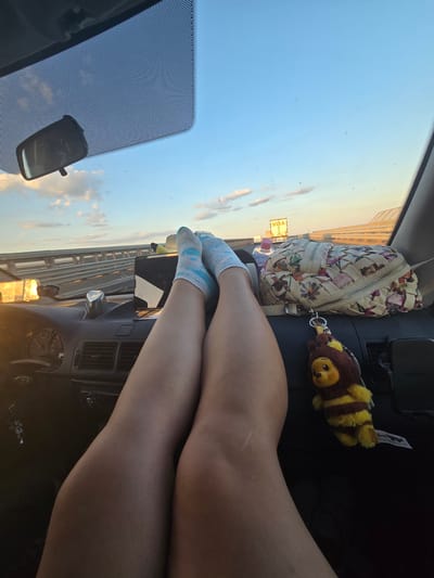 This image depicts the interior of a car, seen from the perspective of someone in the passenger seat, with their legs extended onto the dashboard. The view through the windshield shows a highway with other vehicles and a clear blue sky with some clouds at sunset.
On the dashboard, a patterned handbag with floral designs sits to the right, and a bee-shaped keychain dangles from it. A bottle of water is partially visible behind the handbag. In the foreground, the legs of a person wearing light blue socks with a pattern are positioned. The dashboard itself features car controls, including a steering wheel partially visible on the left, air vents, and a radio. A rearview mirror is attached to the windshield. The overall impression is one of a relaxed moment during a road trip. The specific location in Szeged, Hungary cannot be determined from the image.