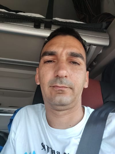 A man is shown in a selfie, taken from the chest up, inside a vehicle. He has short, dark hair, dark eyebrows, and is wearing a white t-shirt. A gray seatbelt is visible across his chest. The background appears to be the interior of a truck cab, with a metallic bar and shelving visible above him. A red and black seat is partially visible behind him. There is no indication of the location within the city of Obersuhl, Germany.