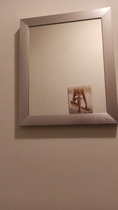 A rectangular mirror with a brushed silver frame hangs on a light beige wall. Reflected in the mirror is a small artwork depicting an abstract figure in a seated or contemplative pose. The artwork is positioned slightly off-center within the reflection. The wall extends to the right, showing subtle color variations and texture. The mirror's shadow is cast on the wall, indicating a light source from the left.