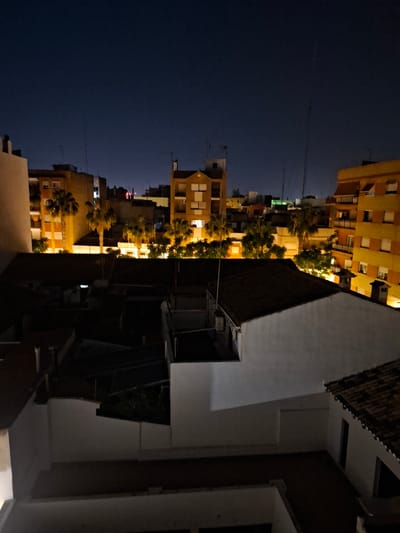 The image shows a night view of buildings in Xirivella, Spain. In the distance, a row of apartment buildings with lit windows stands against a dark sky. Palm trees line the street, adding to the urban landscape. In the foreground, portions of a closer building appear with white walls and dark roof tiles. The overall scene depicts an urban environment at night.