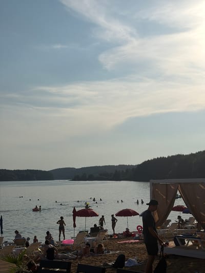 This media file captures a scene at a beach on a sunny day, likely in the late afternoon given the soft, warm light. The setting appears to be a lake or calm sea, with a gentle slope leading into the water. A line of trees densely covers a hill or mountain in the background. The foreground is a sandy beach populated with various beach chairs, umbrellas, and people enjoying the water and sun.
Several individuals are visible in the water, some swimming and others relaxing on inflatable devices. On the beach, people are seated on chairs, some are standing, and others are walking. A prominent red umbrella stands out against the sand and water. The atmosphere seems relaxed and recreational, with a focus on leisure activities.
Notable details include the presence of what appears to be a canopy or tent structure on the right side of the image, casting shade over part of the beach. The sky is mostly clear with some wispy clouds, indicating fair weather. The overall impression is one of a pleasant summer day at a popular waterfront destination. There is no visible text in the image.