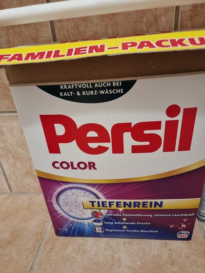 The image shows a close-up of a box of "Persil Color" laundry detergent. The box has a prominent red "Persil" logo and the word "COLOR" underneath. A yellow stripe across the top of the box contains the text "FAMILIEN-PACKU". The bottom portion of the box features a circular design depicting washing powder and the text "TIEFENREIN" which translates to "deep clean". Below this, there are bullet points listing benefits: "Kraftvolle Fleckentfernung, intensive Leuchtkraft" (powerful stain removal, intensive brightness), "Lang anhaltende Frische" (long-lasting freshness), and "Hygienisch frische Maschine" (hygienically fresh machine). A number "90" is visible in a small blue circle, likely indicating the number of washes. The box is placed on a tiled floor with a light brown, textured pattern. The background shows a partial view of a tiled wall. There are no people or animals visible in the image. The lighting is moderate, and the focus is on the product. The environment appears to be indoors, possibly a home or a retail setting, but the location in Pohránov, Czechia, cannot be confirmed from the visual information alone.