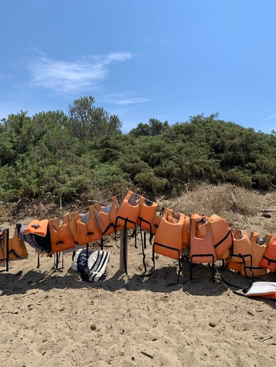 This image depicts a scene on a sandy beach near Orbetello, Italy, likely during daylight hours under a clear blue sky with some wispy clouds. A line of orange life vests is strung up on a simple wooden structure, presumably for drying or storage. The vests are mostly uniformly colored orange with black straps and buckles, and some show signs of wear. To the left, a black and white life vest is lying on the sand. In the background, dense green vegetation, possibly Mediterranean scrub or pine trees, grows on a sandy embankment. The sand itself is strewn with small pieces of driftwood and other debris. The overall impression is one of a recreational area prepared for water activities.