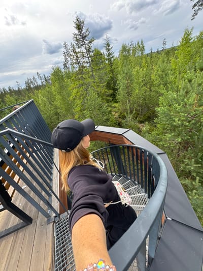An over-the-shoulder shot captures a blonde woman wearing a black baseball cap and black hoodie, looking out at a dense, green forest. She is standing on a circular metal observation deck with wooden planks on the lower level and a grey metal railing. The deck is part of a staircase with metal railings and wooden steps. In the background, a vast expanse of pine trees stretches towards a cloudy sky. A second person, a male, is visible further down the stairs, facing away from the camera. The woman is holding a small, patterned purse in her left hand, and her right arm is extended forward, possibly taking a selfie or holding a phone. The environment suggests an outdoor tourist attraction or viewpoint situated within a natural forest setting, likely in a rural or wilderness area. The location is identified as the city of Peski, Russia.