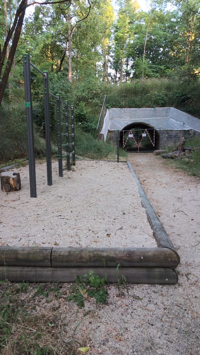 The image depicts an outdoor exercise area in Saint-Apollinaire, France. In the foreground, a sand-covered area features a series of parallel bars for exercise, with a log serving as a bench to the left. Two stacked logs form a border on the left side of the frame.
In the background, a tunnel entrance is visible, covered by a white net. The entrance is blocked by a metal gate with red and white striped caution tape and two warning signs. Surrounding the area are dense green trees and vegetation. The scene appears to be a public park or recreational space.