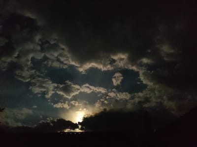 The image shows a dramatic nighttime sky over the city of Qargaly, Kazakhstan, with the moon partially obscured by dark, heavy clouds. The moon's light creates bright, luminous edges on the clouds, giving them a dramatic and almost ethereal appearance. In the foreground, a dark silhouette of a treeline or the edge of a building is visible against the illuminated sky. The overall impression is one of a stormy night with a powerful display of celestial light.