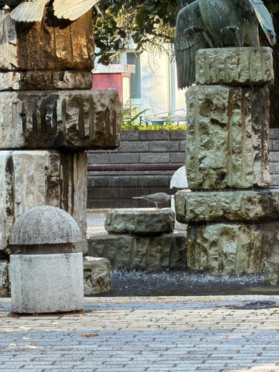 This image depicts a close-up view of an outdoor sculpture in Tiszaszentmárton, Hungary. The sculpture is made of rough, textured stone blocks, some of which appear to be weathered and stained. There are also elements that suggest a fountain or water feature, with water running down some of the stone surfaces, creating a glistening effect.
A dove is perched on one of the stone ledges, its feathers a soft gray. In the background, a building with windows and a red door is visible, partially obscured by foliage. The lighting suggests it is daytime, with sunlight illuminating parts of the scene and casting shadows. The overall impression is one of an artistic installation in a natural setting, possibly a garden or a public space. The texture of the stone and the presence of the bird add a sense of organic detail to the man-made structure.
