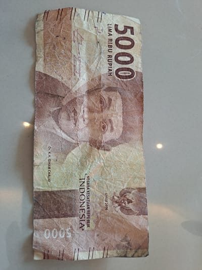 The image shows a crumpled 5000 Indonesian Rupiah banknote lying on a light-colored, textured surface. The banknote features a portrait of a man, the denomination "5000", and text in Indonesian. The banknote has visible creases and wear. The background surface appears to be a countertop or table. The image does not provide any information about the city of Bireuenmeunasahdayah, Indonesia.