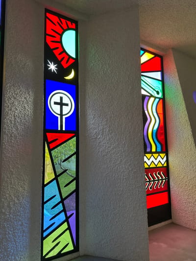 The image depicts an interior view of a building, focusing on two stained glass windows. A heavily textured, off-white wall column separates the two windows.
The left, more prominent window is composed of multiple vertical panels. The uppermost panel features a red background with a radiant, light blue sun on the right, and a white star and yellow crescent moon positioned below the sun on a dark segment. Below this, a blue panel displays a white circular symbol containing a black cross, atop a white stylized staff. The lowest section of this window features an abstract geometric design composed of colored glass in shades of red, yellow, green, blue, and purple, with black leading lines; through some green and light blue sections, an outdoor view of a tree and green foliage is visible.
The right window, partially obscured by the wall, also features abstract stained glass designs. Visible elements include red, yellow, and green geometric shapes in its upper part, followed by a prominent wavy pattern in purple, yellow, light blue, and red. Below this, a white zig-zag pattern on a red background is present, followed by repetitive, stylized red and white shapes on a dark background.
Natural light passes through both windows, casting colored reflections, primarily green, purple, and red, onto the textured off-white walls and light grey floor. No people, subjects, or interactions are present in the scene.
This location is within the Galil Elyon Regional Council, Israel.