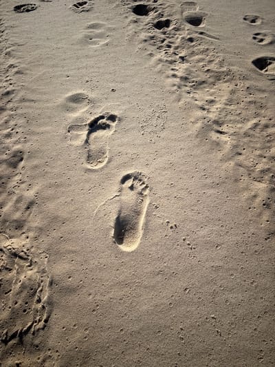 The image depicts footprints in sand. There are two clear footprints from an individual in the central and lower-left portion of the image. Additional footprints are discernible in the upper-left area of the image. The sand exhibits variations in texture and density with small holes throughout the flat areas. No people or other subjects/objects are immediately visible in the frame beyond the sand and its markings.
Location: This is likely a beach in Ciudad de la Costa, Uruguay given its sandy environment.