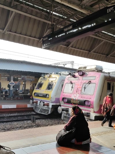 The scene depicts a railway station platform in Bardhaman, India, during daylight hours. In the immediate foreground, a woman wearing a dark top, patterned headscarf, and light-colored skirt is seated on the tiled platform, facing left. A dark bag rests beside her. Directly behind her, two multi-colored Electric Multiple Unit (EMU) train cars are stationary on parallel tracks. The train car on the left is predominantly yellow and white with purple accents, displaying "ICF" and the number "160439". The train car to its right is predominantly pink/magenta and white, also marked "ICF" and bearing the number "258357". Both train cars feature front grilles, headlights, and windshield wipers.
On the platform opposite the trains, several individuals are visible, some seated on benches and others standing. A blue and white station sign is present on this distant platform. A person wearing a red patterned shawl walks past the pink train car on the right side of the frame. Overhead, a large digital information board hangs from the station's corrugated metal roof structure, displaying "TRAIN NO." and "TRAIN NAME" in white text against a dark background, with illuminated dots below indicating active display. The sky is bright and clear, with overhead electrical lines and support structures visible.