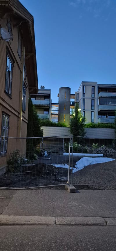 In Lillehammer, Norway, on a construction site, a wire mesh fence separates a sidewalk from a cleared area. The area behind the fence contains piles of gravel, white plastic sheeting, and some debris, suggesting ongoing renovation or construction. A cylindrical tower structure with illuminated windows is visible in the background, part of a modern apartment building. To the left, an older wooden house with visible architectural details and a satellite dish stands. The sky is a clear, deep blue, indicating early evening or twilight.