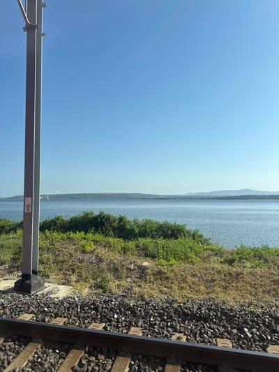 An image taken from a train window shows a view of a body of water. In the foreground, train tracks run horizontally across the bottom of the frame. Beside the tracks, a tall, grey metal pole stands vertically on the left side of the image. A small white sign with black text and numbers "420" and "18" is attached to the pole. Beyond the pole and tracks, there is a slope covered in dry grass and green bushes. The water is calm and blue, extending to the horizon. In the distance, a hilly landscape with some faint structures is visible. The sky is clear and bright blue, with a single faint contrail from an airplane. The scene is bathed in natural daylight.