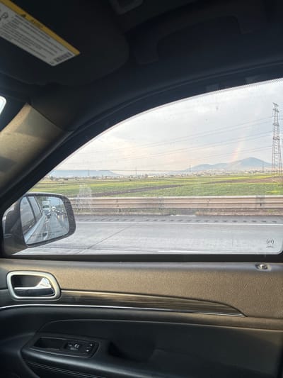 The image depicts a view from inside a vehicle, likely a car, looking out through the passenger side window and the side mirror. The window shows a landscape featuring a highway with multiple lanes, next to it is an agricultural area. The landscape also includes buildings and mountains in the distance. A rainbow is faintly visible in the sky. Power lines are seen stretching across the landscape. The side mirror reflects the highway with other vehicles. The interior of the car is visible, including the door panel, window control, and a portion of the sun visor.

The location is indicated to be Coyotepec, Mexico, based on the metadata.