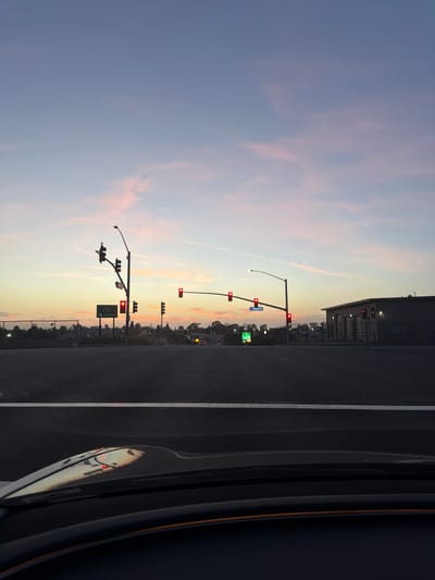 The image depicts a sunset scene at a street intersection in Newport Beach, California, United States. The sky is a gradient of pale blue and pink hues, with thin, wispy clouds. In the foreground, the dashboard of a vehicle is visible, reflecting the sky and traffic lights.
The intersection features multiple traffic signals, all illuminated red, indicating that vehicles must stop. A green highway sign with the number "73" and the word "SOUTH" is visible on the left side of the road. A blue street sign reading "Campus Dr" is also present. In the background, buildings and streetlights are silhouetted against the twilight sky. A chain-link fence runs along the side of the road. The road surface is asphalt, with a white lane marker clearly visible. No people are visible in the frame.