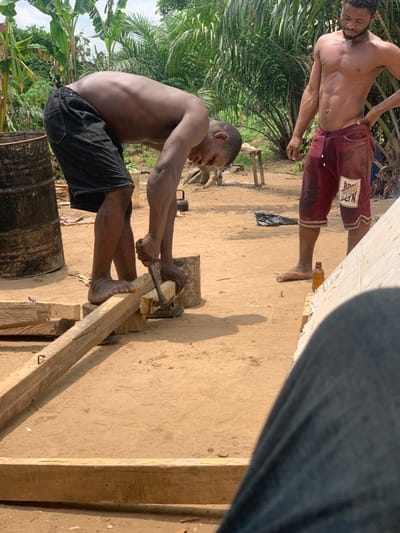 The image captures two men in an outdoor setting. One man, shirtless, is wearing red shorts with the inscription "Brown Bell" visible. He stands to the left of the frame and looks towards the center. The other man, also shirtless, is wearing black shorts. He is positioned in the center of the frame, crouching down and using a metal tool to work on a wooden plank. The plank is resting on two other pieces of wood, and there is a piece of wood beneath it. The ground appears to be loose dirt, and there are various pieces of wood scattered around. Behind the men, there are lush green plants growing in the background. The location is Egbeda, Nigeria, based on the provided coordinates (latitude: 7.12, longitude: 5.362).