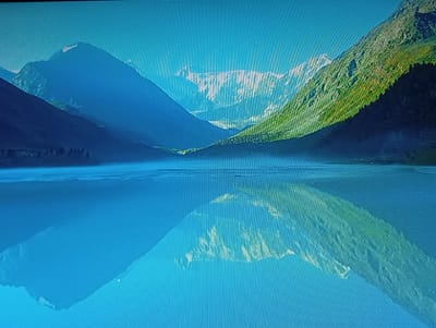 The image depicts a serene mountain landscape reflected in a still body of water. Majestic, blue-hued mountains dominate the background, with a lush, green mountainside on the right. The sky above is a clear, bright blue. The reflection in the water perfectly mirrors the mountains, creating a symmetrical and tranquil scene. There are no people or man-made structures visible in the image, suggesting a remote natural setting. The scene is likely a picturesque vista, possibly a lake or fjord in a mountainous region. This image is not related to Olișcani, Moldova, as it portrays a mountainous, natural environment.
