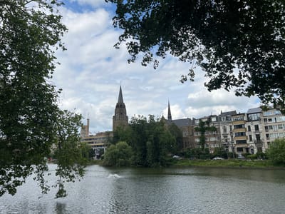 A serene body of water, likely a canal or lake, fills the foreground and mid-ground of the image. The water's surface is rippled, reflecting the overcast sky and surrounding greenery. In the distance, a complex of buildings lines the opposite bank. Prominent among these are two church spires, one taller than the other, constructed of brick and stone. Adjacent to the churches are multi-story residential and commercial buildings with varied architectural styles. Green trees and vegetation are abundant throughout the scene, with branches from foreground trees partially obscuring the view. A fountain with white spray is visible in the water to the left of center. The sky above is a mix of white clouds and blue patches. The scene appears to be the Ixelles Ponds in Ixelles, Belgium, a popular urban park area.