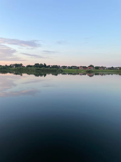 The image depicts a serene landscape at what appears to be either sunrise or sunset, with a tranquil body of water in the foreground and a tree-lined horizon with scattered houses in the background. The sky is a gradient of soft blues and pinks, with scattered clouds. The water perfectly reflects the sky and the distant landscape, creating a symmetrical composition. The houses appear to be residential, varying in architectural style and color, and are nestled amongst lush green trees. The overall atmosphere is peaceful and idyllic. The location is identified as Sovetsk, Russia.