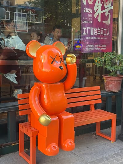 An oversized, glossy orange vinyl figure resembling a stylized bear, with gold accents on its hands and ears, is seated on an orange slatted bench outside a storefront. The figure has black "X" marks for eyes. Behind the figure and bench, through a large glass window, several people are visible. A woman with dark hair is seated on the left, looking to her left, with a white plastic bag on the table in front of her. To her right, a young girl with dark hair tied back is looking towards the right. Further right, a man with short dark hair is seated, looking down. A poster with Chinese characters and "JOIN US" is affixed to the glass window. To the right of the bench, a potted plant with green leaves and small orange flowers is situated. The scene appears to be in an urban setting, potentially a commercial area in 幸福乡, China. The ground is paved with grey tiles.