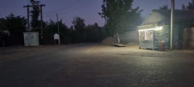 This image depicts a dimly lit street scene in the city of Havaskor, Tajikistan, during twilight or night. The foreground shows a paved road with a slight sheen, suggesting it might be damp or freshly paved. To the right, a small, brightly lit kiosk or shop is visible, stocked with various items displayed behind glass. A large orange trash bin overflowing with garbage is situated next to the kiosk. In the midground, a pile of dirt and some wooden planks are present. Further to the left, there's a utility box or small structure with a blue-green hue, accompanied by a wooden utility pole with electrical wires extending overhead. Trees and foliage are scattered throughout the background, with a larger tree prominently positioned near the dirt pile and kiosk. The sky is a deep blue, indicating low light conditions. There are no people visible in the image.