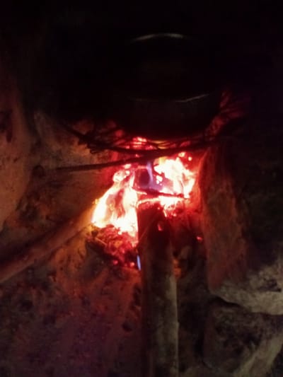 The image depicts a rustic cooking scene, likely at night or in a dimly lit enclosure, given the profound darkness surrounding the central light source. The primary subject is a wood-burning fire, blazing with bright orange, red, and yellow flames, and glowing embers at its base.
Above the fire, a dark, round cooking pot or container is visible, resting on what appears to be a simple metal grate or support structure, indicating that something is being cooked or heated. Fuel for the fire, consisting of various pieces of wood or branches, is present, with some directly feeding the flames and others laid around. The fire is contained within a rough, earthy or stone structure, possibly a traditional three-stone fire or a simple mud stove, designed to hold the heat and support the cooking vessel. The texture of these walls appears coarse and natural.
No people are visible in the frame. The overall atmosphere conveyed is one of warmth and basic sustenance, typical of traditional cooking methods. There is no visible text. The setting is consistent with a rural or semi-rural environment in Mugumo-ini ward, Kenya, where such traditional cooking setups are common.