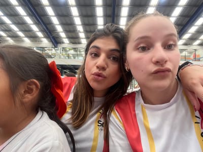 Three young women are posing for a selfie indoors, likely at a school event in Tlaxcalancingo, Mexico. The two women in the center and right are positioned closely, making a face for the camera. They are wearing matching white t-shirts with red and gold stripes and a logo with the letter "S". The woman on the left has her back towards the camera, only partially in view with a red bow in her hair. The environment appears to be a gymnasium or large indoor space, characterized by a high ceiling with metal support beams and lighting fixtures.