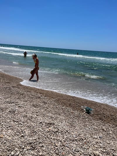A sunny day at Ga'ash Beach in Israel. The foreground is a pebbly beach with a plastic water bottle lying on its side near the water's edge. In the middle ground, a woman in a yellow bikini stands in the shallow water, holding a straw hat. Further out in the ocean, several people are visible wading in the turquoise water with white waves breaking. The background features a clear, bright blue sky.