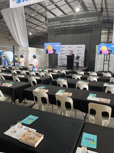 The image shows a large indoor space set up for a seminar on sake.  The scene is taken from a seated position in the audience, facing the front.  The location is a large warehouse or convention center located at latitude: -27.439, longitude: 153.075 in Hamilton, Australia.  Black tablecloths cover tables, set up in rows with white chairs.  A few people are standing, but most are seated.  Three screens on the stage display the words "SAKE SEMINAR," with two people standing in front of each screen, one is giving a presentation.  There are some empty glasses and other objects on the tables.  A large white banner hangs from the ceiling.  The room has an industrial feel, with exposed rafters and steel girders.