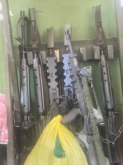 The image shows a rack of rifles, likely stored indoors. The rifles are dark in color and appear to be made of metal and synthetic materials. They are positioned vertically, with straps attached to some of them. A yellow plastic bag is visible in the foreground, partially obscuring the rifles. The background wall is painted green. The scene appears to be a storage area for firearms.