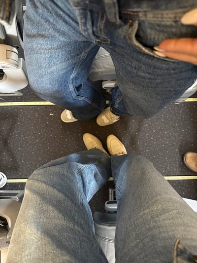 A top-down shot showcases the legroom between four airplane seats. Occupants wear jeans and casual footwear. On the left, a person sports sneakers with a white base, while the person across wears beige loafers. A third person has on tan leather shoes. The seats are gray and the carpet is dark with small, light-colored speckles, and a thin yellow aisle marker is visible.