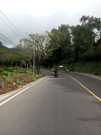 The image depicts a curved asphalt road in Saing Ta Mawt Lay, Myanmar, on an overcast day. The road, running from the lower left to the upper right, features a solid white line marking the left shoulder and a solid yellow line indicating no passing in the center.
On the left side of the road, an unpaved embankment shows exposed dirt, small grey concrete blocks, and green netting, suggesting recent ground work or erosion control. Beyond this, scattered trees with varying foliage, some appearing deciduous, line the road. A tall grey metal streetlight pole stands prominently on this side. Multiple dark power lines or cables are strung across the upper left quadrant of the sky. In the distant background on the left, hazy green hills are visible beneath the cloudy sky.
The right side of the road is bordered by a steep, densely vegetated slope covered in various green trees and bushes, with a metal guardrail partially visible along the edge. A white rectangular sign with illegible text is affixed to the slope further down the road.
Three motorcycles are traveling on the road, moving away from the viewer. The closest, positioned centrally, is a dark-colored motorcycle carrying two individuals in dark clothing and helmets. Ahead and to its left is a lighter-colored scooter with a single rider in a light helmet and jacket. Further ahead on the right, a blue or light-colored scooter with a single rider is visible. In the far background, a dark-colored vehicle is partially visible near a road bend on the left. The sky is uniformly light grey and cloudy.
