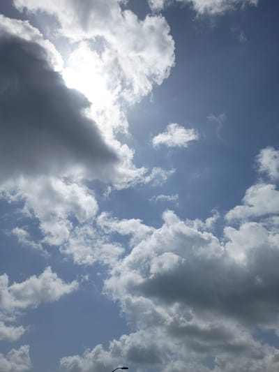 The image shows a sky filled with a mix of dark and light clouds. The sun is partially obscured by a large, bright white cloud formation in the upper left quadrant, casting a bright glow and rays of light. The sky itself is a clear blue, with smaller, wispy clouds scattered throughout. In the very bottom center, a portion of a street lamp is visible, with its curved arm extending upward. The scene appears to be an outdoor view, possibly from street level, in Nicholasville, Kentucky, USA, given the reference to the location.