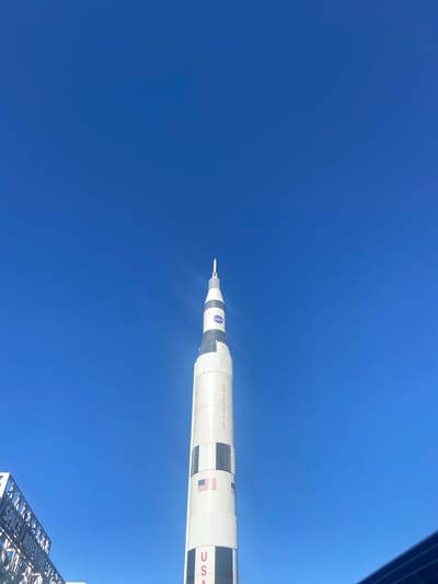 The primary subject of the image is a massive, white and black multi-stage rocket, resembling a Saturn V, seen against a clear, deep blue sky. Although the original image is oriented vertically and then rotated 90 degrees clockwise, the rocket itself is an upright structure. Its pointed nose cone is visible near the top, while its main body extends downwards, occupying the right side of the frame.
Prominent text and logos are visible on the rocket's exterior: the classic circular blue "NASA" emblem is near the top section, followed by the words "UNITED STATES" written vertically. Further down, the letters "USA" are partially visible. The sky is a uniform, intense blue, devoid of clouds, indicating bright, sunny daylight. A subtle lens flare or sun glint can be observed near the rocket's tip, hinting at the sun's position.
In the bottom right corner, a white architectural structure with an open, lattice design is partially visible. Etched into this structure are the words "SPACE ADVENTURE," clearly identifying the location as a space-themed attraction. No individuals are present in the frame, allowing the monumental scale of the rocket to dominate the scene. This outdoor display is situated at the Space Adventure museum in Canela, Brazil, presenting an impressive homage to space exploration under a vast, open sky. The overall atmosphere conveys awe and technological achievement.