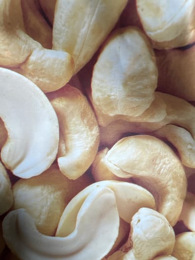 This is a close-up, overhead shot of a pile of raw, whole cashews. The nuts are light beige to pale yellow in color and have a slightly textured surface. They are irregularly shaped, with some appearing curved or kidney-bean-like, while others are more oblong. Some cashews are whole, showing their distinctive curved shape, while others appear to be broken or partially obscured. The background is a warm, muted brown, suggesting the shallow depth of field. There is no visible text. The image is well-lit, with soft light that highlights the textures and subtle color variations of the nuts. The focus is sharp on the cashews in the foreground, with a slight blur towards the back, creating a sense of depth and emphasizing the subjects. The overall impression is one of natural, unprocessed food.