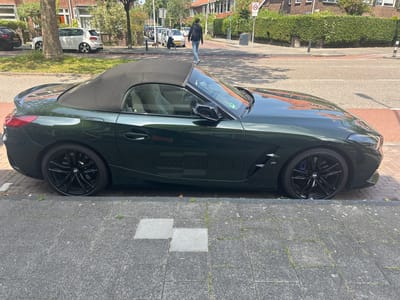 A dark green convertible BMW Z4 is parked on a paved street in Leiden, Netherlands, alongside a sidewalk with grey paving stones and two lighter grey square inlays. The car's black soft top is up. The street is lined with parked cars, residential buildings with brick facades and tiled roofs, and dense green hedges. A person in a black jacket and blue jeans is walking across the street in the background, passing a round 30 km/h speed limit sign. The sun is casting shadows on the street and pavement.