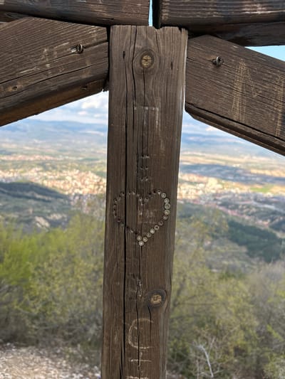 This image shows a weathered wooden crossbeam structure, likely part of an overlook or viewing platform. The crossbeam is decorated with a heart shape formed by numerous small, round, metallic objects, possibly screws or decorative studs. There are also carved inscriptions on the wood, with the letters "OH" faintly visible. The backdrop features a panoramic view of a city nestled in a valley surrounded by green hills and trees. Based on the prompt, the city is Delchevo, Bulgaria. The sky is partly visible in the upper left corner.