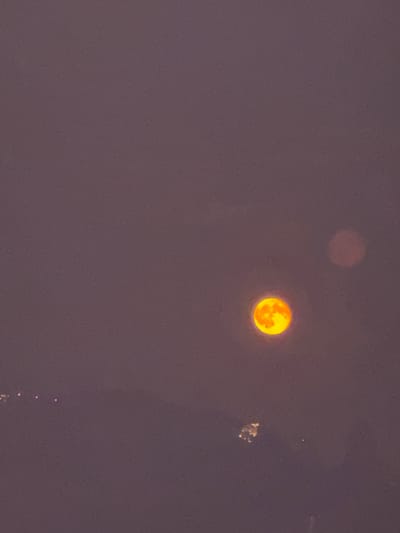 The image shows a low-light view of the night sky over the area of Balerna, Switzerland. A prominent bright orange full moon is visible in the center-right of the frame, with visible surface details. To the upper right of the moon, a fainter, circular reddish-brown blur is present, possibly another celestial object or lens flare. In the lower left of the image, a cluster of small, distant lights is visible, suggesting urban or populated areas. In the lower center, a scattered group of faint lights appears to be on the ground or water. The overall sky color is a muted purple-grey, indicating twilight or a heavily overcast sky.