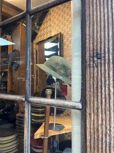 The image displays a detailed view into Laird Hatters, a hat shop located on Turl Street in Oxford, United Kingdom, captured through a multi-paned window with a dark, weathered wooden frame.
In the center, a green tweed bucket hat with subtle orange and yellow plaid detailing and small gold-colored studs is prominently displayed on a silver metal stand, positioned on a light wooden shelf. To the left, multiple stacks of various hats, including fedoras and panamas in different shades, are visible on lower shelves and within a dark wooden cabinet.
The interior background reveals light-colored wallpaper adorned with a repeating serpentine or wave pattern in red, blue, and black lines. A large mirror with an ornate dark frame reflects parts of the ceiling, including dark beams, and bright interior lights. Within the reflection or visible deeper in the shop, a person wearing dark clothing, potentially a hoodie or beanie, is observed looking downwards, seemingly engaged in work or inspecting an object on a surface. A red tag with "Laird Hatters" text is visible hanging. A brass-colored fitting is attached to a wooden support column in the lower center foreground.
The right side of the image features a vertical section of an external, weathered dark wooden facade or door frame, characterized by deep, prominent vertical grooves.