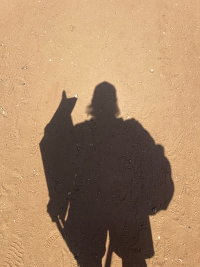An overhead shot shows the dark shadow of a person standing on sandy ground in the city of Gaash, Israel. The person's shadow is cast in sharp relief by the sunlight, with the head appearing to wear a hat and the left arm raised in a peace sign. A backpack or bag is visible on the right side of the shadow's back. The sandy ground displays tire tracks, footprints, and small stones and pebbles scattered across its surface.