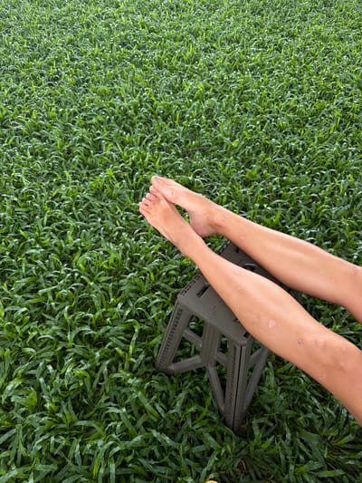 In Tambon San Phi Suea, Thailand, the image captures a close-up view of two bare feet and lower legs resting on a dark green, foldable plastic stool. The stool is positioned on a dense, vibrant green lawn, likely a grassy area. The legs exhibit visible skin variations and minor marks. The lighting suggests an outdoor environment, possibly during daylight hours, with natural light illuminating the scene. The overall impression is one of relaxed outdoor activity.